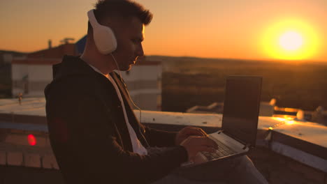 A-man-freelancer-in-headphones-standing-on-the-roof-at-sunset-writes-on-the-keyboard-code-pages.-Little-business.-Listen-to-music-and-work-at-the-computer-enjoying-the-beautiful-view-from-the-roof.